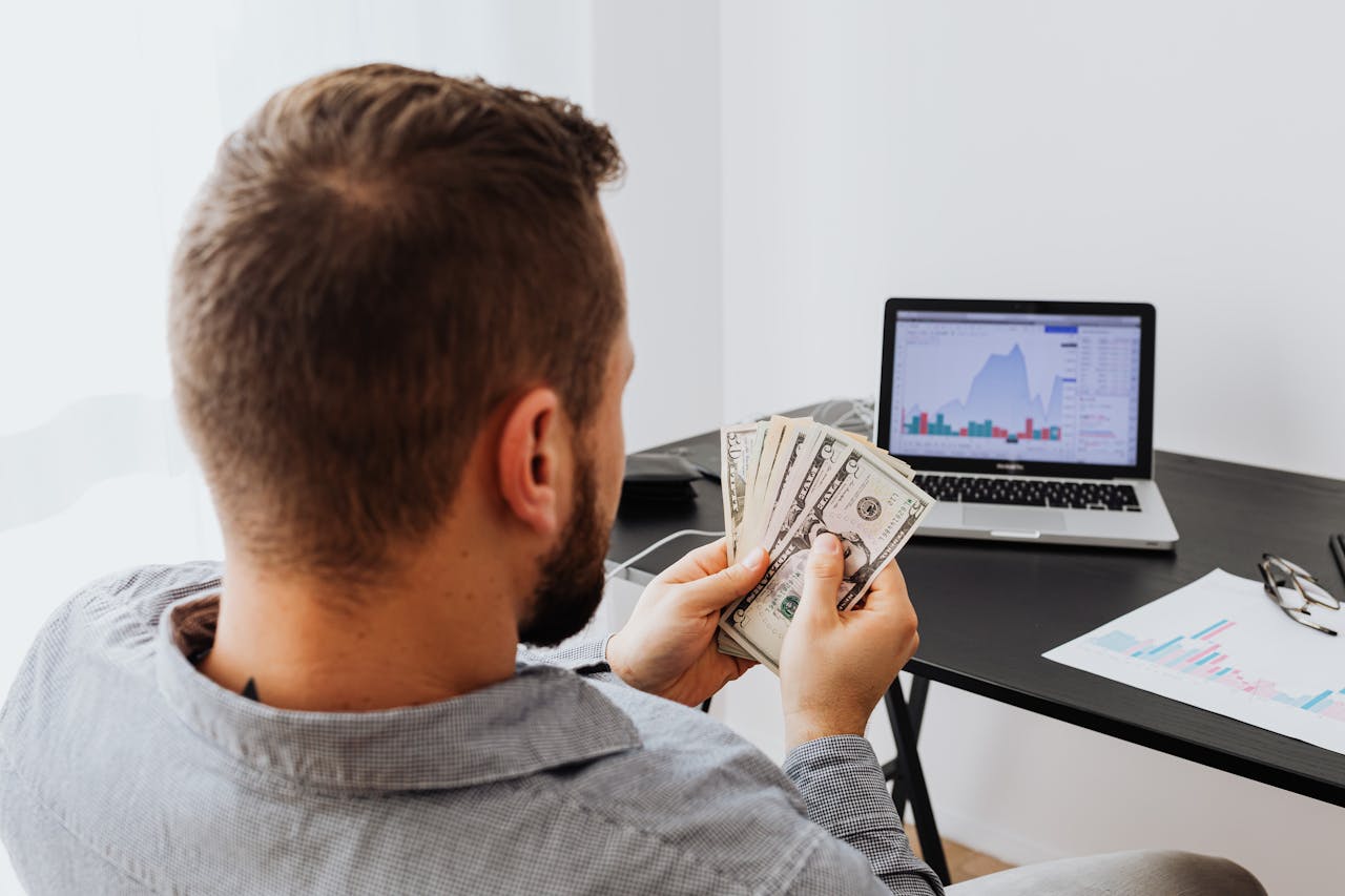 Man counting money at a desk with a laptop showing financial data.