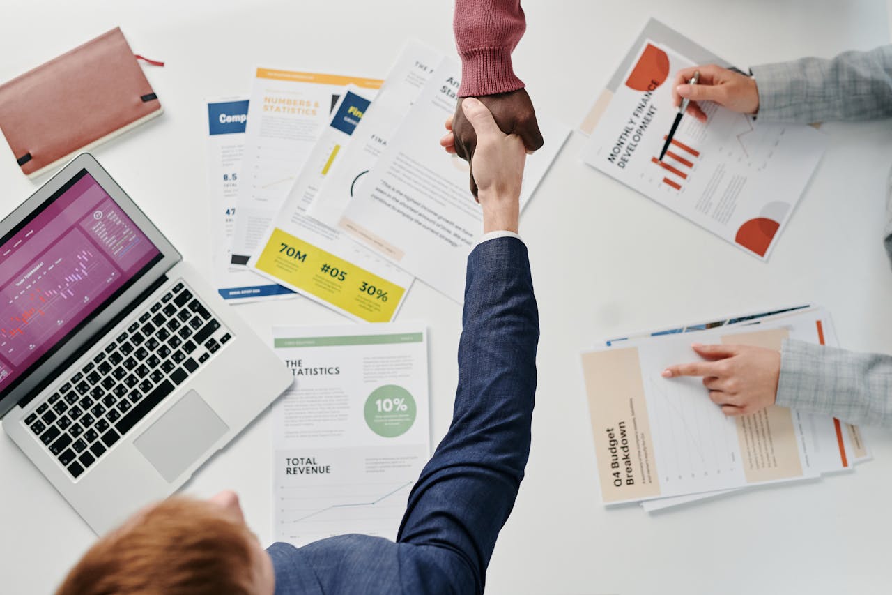 Offerings Business professionals engaging in a handshake over a desk filled with financial documents and a laptop.