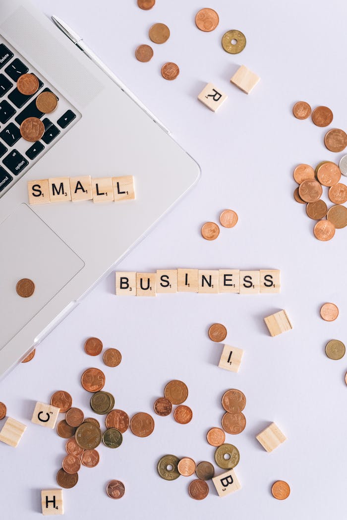 Flat lay of coins, Scrabble tiles spelling 'small business', and laptop, perfect for finance or entrepreneurship themes.
