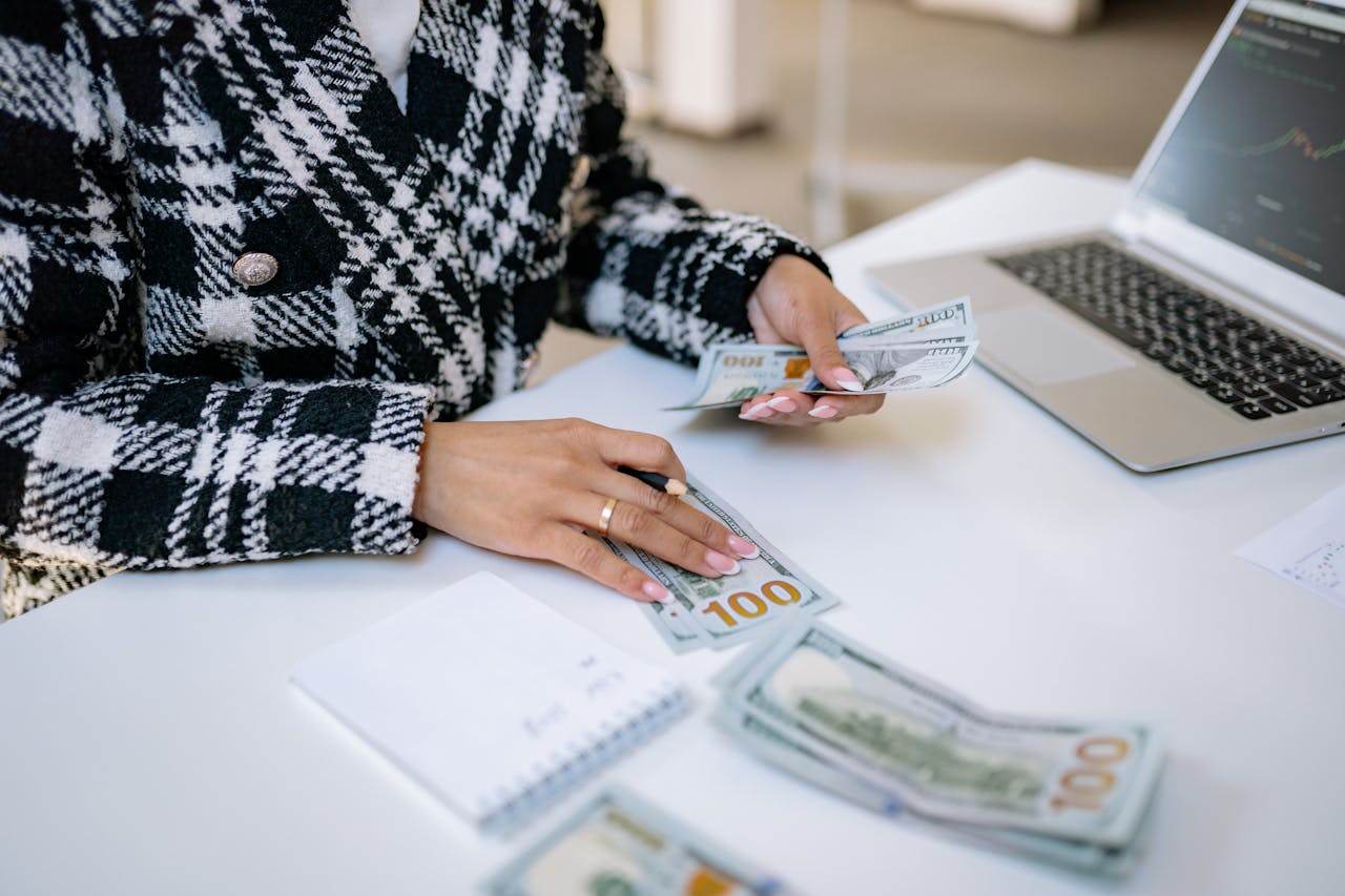About Close-up of a woman counting hundred-dollar bills at a desk with a laptop, focusing on finance and work.